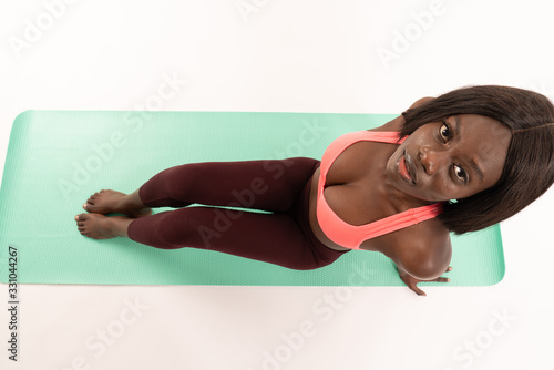 Top view photo of young african girl in stylish sportswear who sitting om the yoga mat and looking at the camera