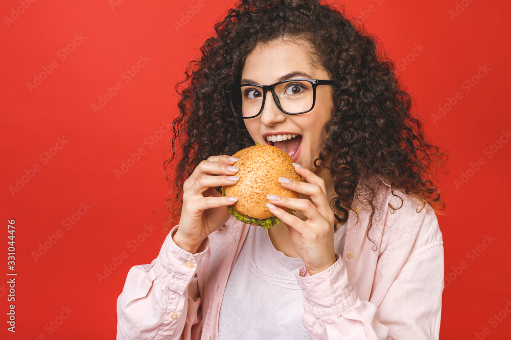 Portrait of young curly beautiful hungry woman eating burger. Isolated portrait of student with fast food over red background. Diet concept. Portrait of young curly beautiful hungry woman eating burger. Isolated portrait of student with fast food over red background. Diet concept.