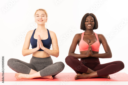 Happy international girls in sportswear. Concept of healthy life and balance between body and mental development. Gorgeous young female's sitting on the red yoga mat closing their eyes, holding hands