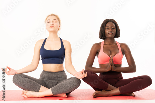 International girls in sportswear sitting in lotus position, african and european female's meditating with closed eyes while sitting on a yoga mat