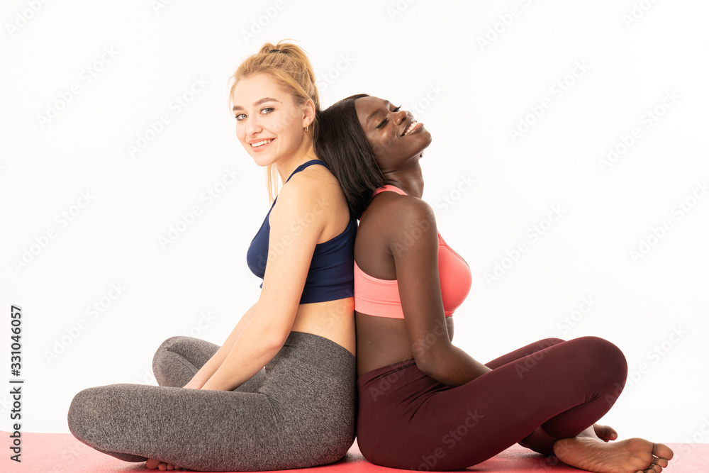 Obraz premium Relaxing together. Two beautiful international young women in sports clothing sitting back to back at lotus position. Bottom view