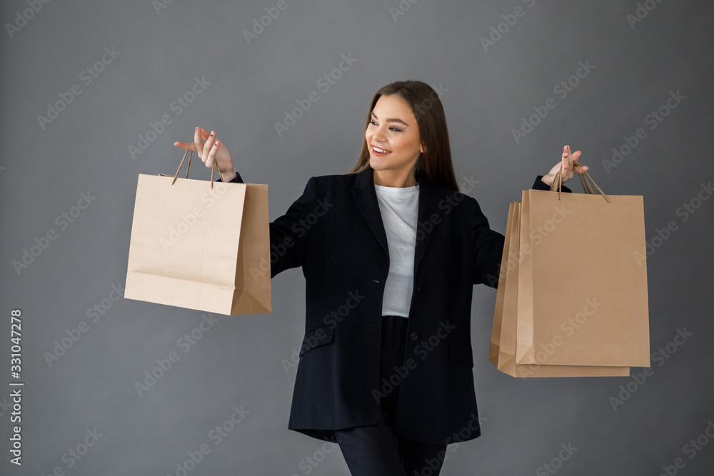Young pretty girl in business style clothes stands on gray background with paper bags in hands. Business style Young pretty girl in business style clothes stands on gray background with paper bags in hands. Business style