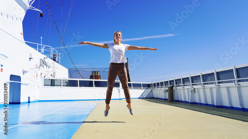 Beautiful sporty girl jumps up on the deck of a cruise ship on a background of blue clear sky. A happy girl smiles on vacation while traveling, jumped high and spread her arms to the sides. © Artem