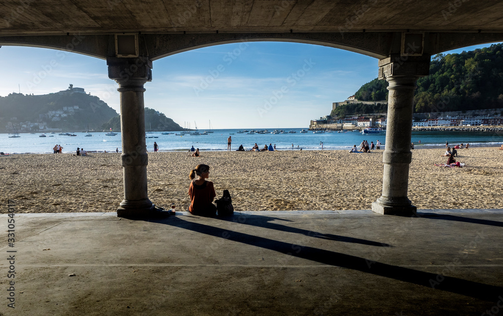 Fototapeta premium Chica joven sentada junto a dos columnas de piedra al borde de la Playa de La Concha de San Sebastian