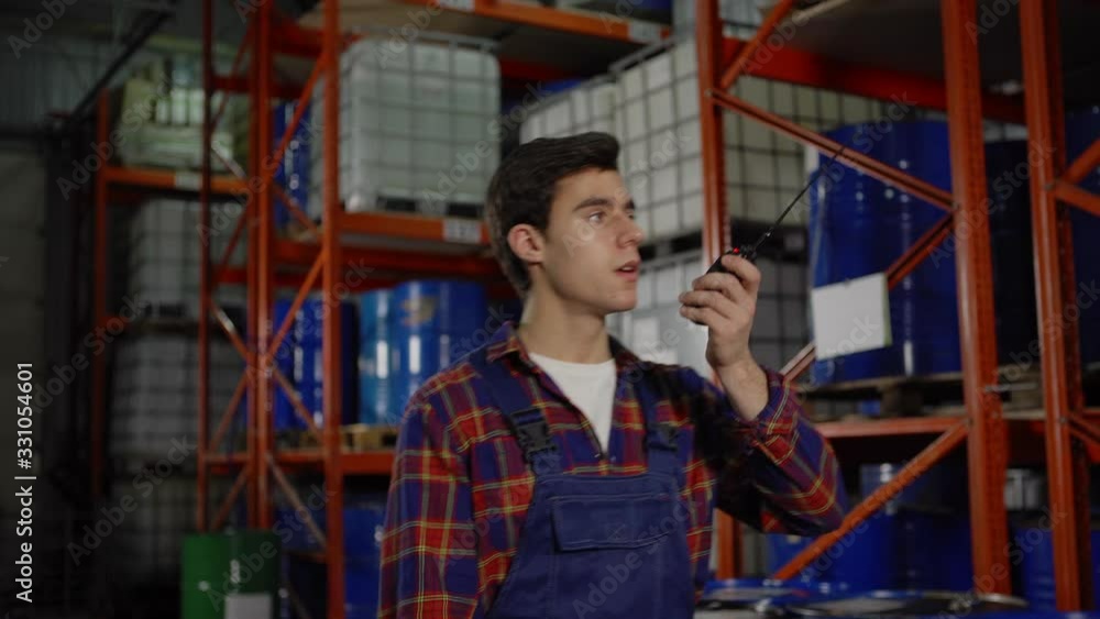 Tracking medium shot of middle engineer in white coat walking in factory storage while young warehouse worker talking on walkie-talkie passing by racks with steel barrels and containers Tracking medium shot of middle engineer in white coat walking in factory storage while young warehouse worker talking on walkie-talkie passing by racks with steel barrels and containers