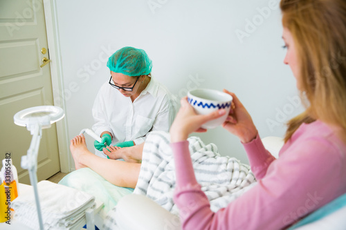 Woman relaxing in chair with cup of coffee, having foot treatment in spa salon. Professional medical pedicure specialist in protective mask using special electric lathe and tools. 
