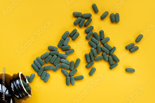 Pile of Green spirulina pills dropped out of the jar on yellow background.Spirulina tablets background.Top view, flatlay.Pile of tablets. Spirulina dietary supplement, alternative medicine photo