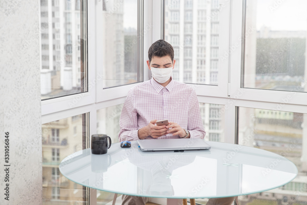 A guy in a protective face mask is typing on the phone and working on a computer from home during a virus epidemic. Quarantine during viral infection A guy in a protective face mask is typing on the phone and working on a computer from home during a virus epidemic. Quarantine during viral infection