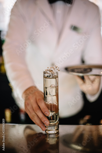 Bartender in a white suite and black bow tie holding on his hand gin and tonic cocktail in a highball glass decorated with a bunch of dried flowers. Smooth image with shallow depth of field.