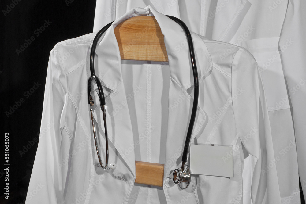 White medical gowns hang on hangers. Clothes with stethoscope on black background. White medical gowns hang on hangers. Clothes with stethoscope on black background.