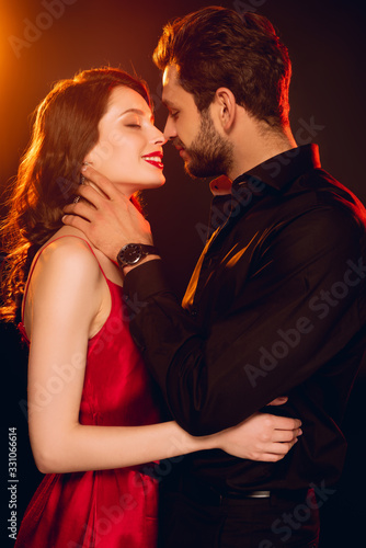 Side view of elegant man touching neck of beautiful smiling girl on black background with lighting