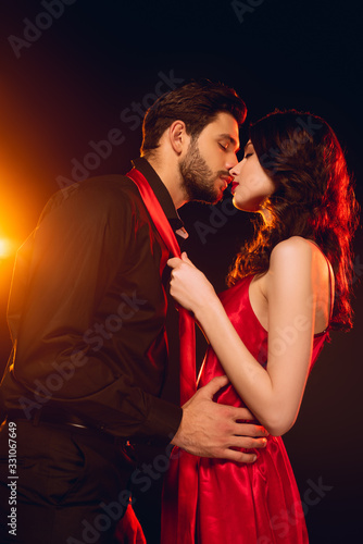 Side view of girl in red dress kissing and holding tie of handsome man on black background with lighting
