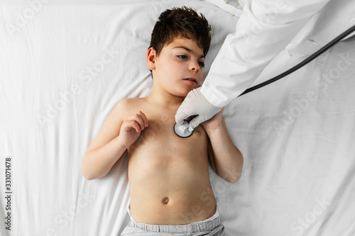 un niño en una cama es auscultado por una doctora.  tiene el estetoscopio (fonendo) en los pulmones del niño. la doctora lleva guantes de latex blancos y bata blanca. photo