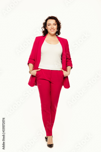 Full length of smiling businesswoman in red suit posing while stands holding hands in pockets and looking at camera isolated over white background.