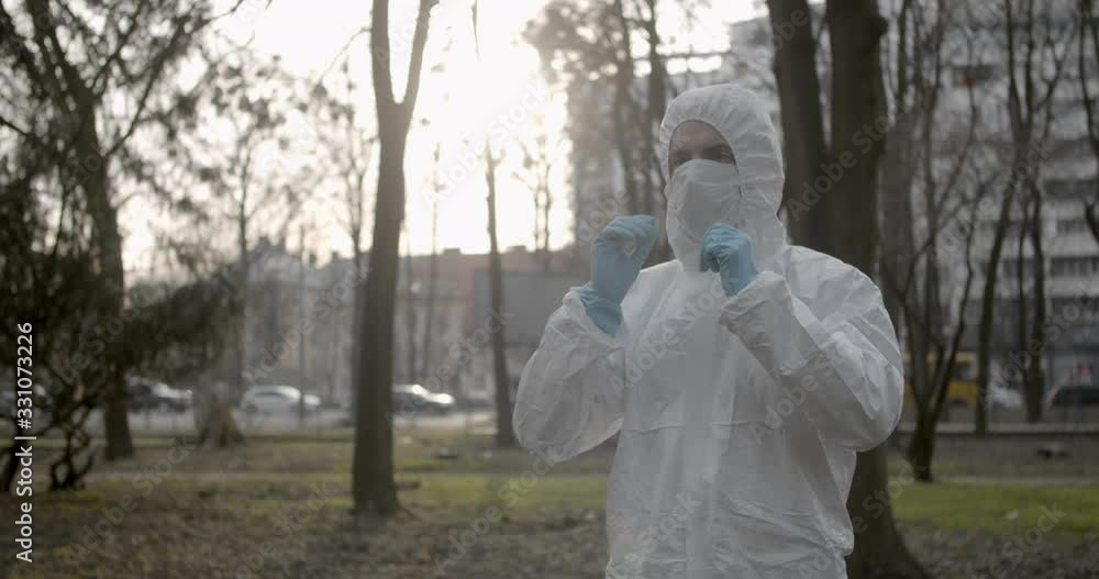 Portrait of male scientist in protective suit wearing a glove and adjusting his mask in daytime park in pandemic coronavirus quarantine time as cars and pedestrians pass by 4K Portrait of male scientist in protective suit wearing a glove and adjusting his mask in daytime park in pandemic coronavirus quarantine time as cars and pedestrians pass by 4K
