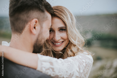 Smiling bride and groom spending time together. Posing on the mountain hills background. Dressed in white dress beautiful blonde caucasian bride and handsome groom. Hugs, kissess and enjoy the company