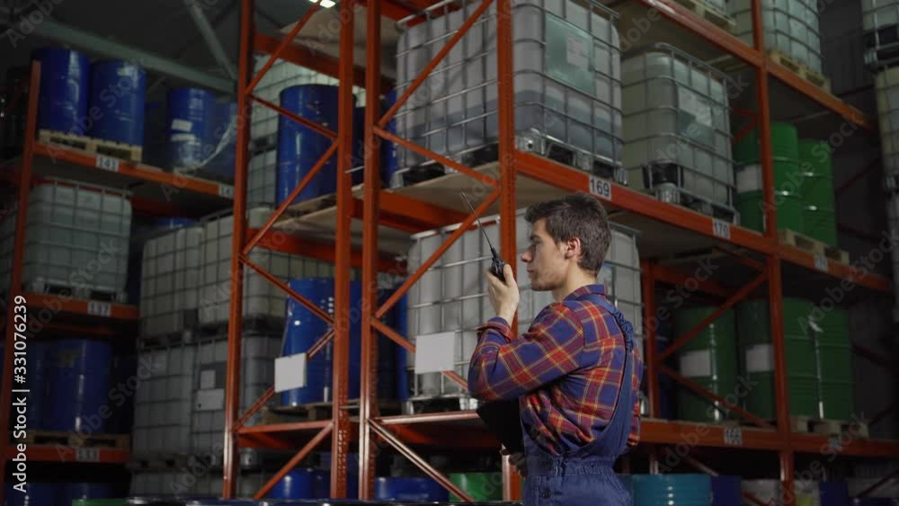 Side view tracking shot of young warehouse worker in uniform talking on walkie-talkie standing by racks with steel barrels and containers in factory storage Side view tracking shot of young warehouse worker in uniform talking on walkie-talkie standing by racks with steel barrels and containers in factory storage