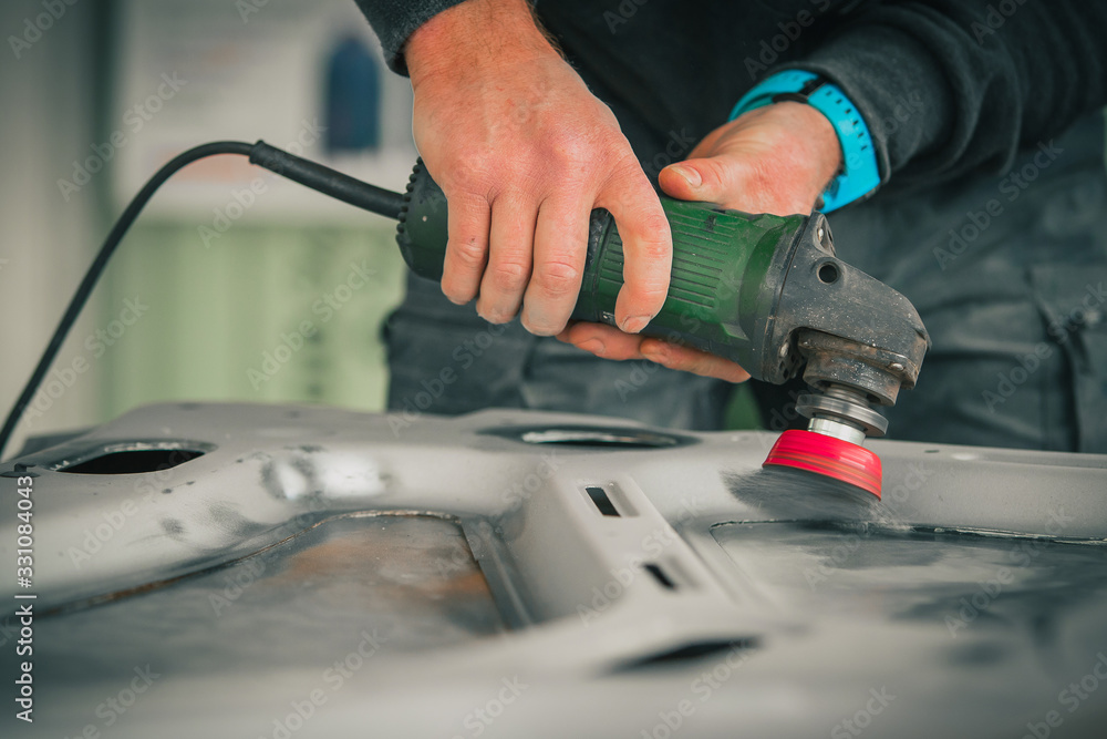 Young service man is using an angle grinder with a wire brush to clean the underside of an old vintage car about to be restored Young service man is using an angle grinder with a wire brush to clean the underside of an old vintage car about to be restored