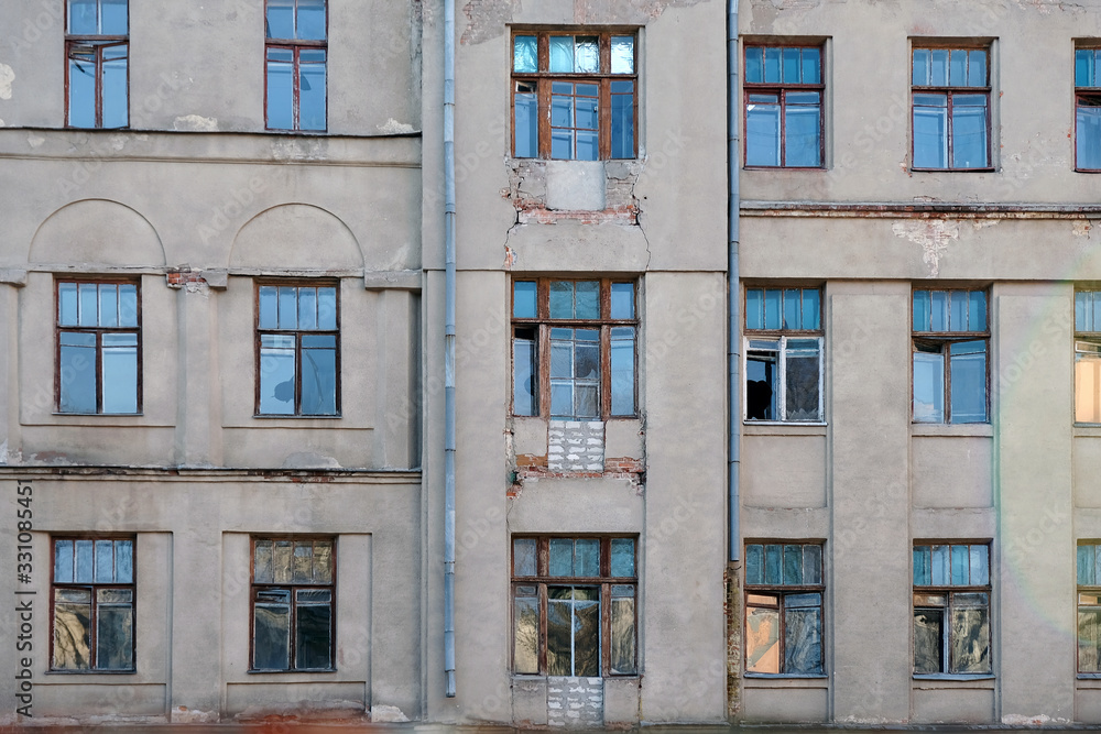 Old building facade with broken windows. Gray multi-storey building with windows of different sizes. Abandoned building with wooden frames for windows.