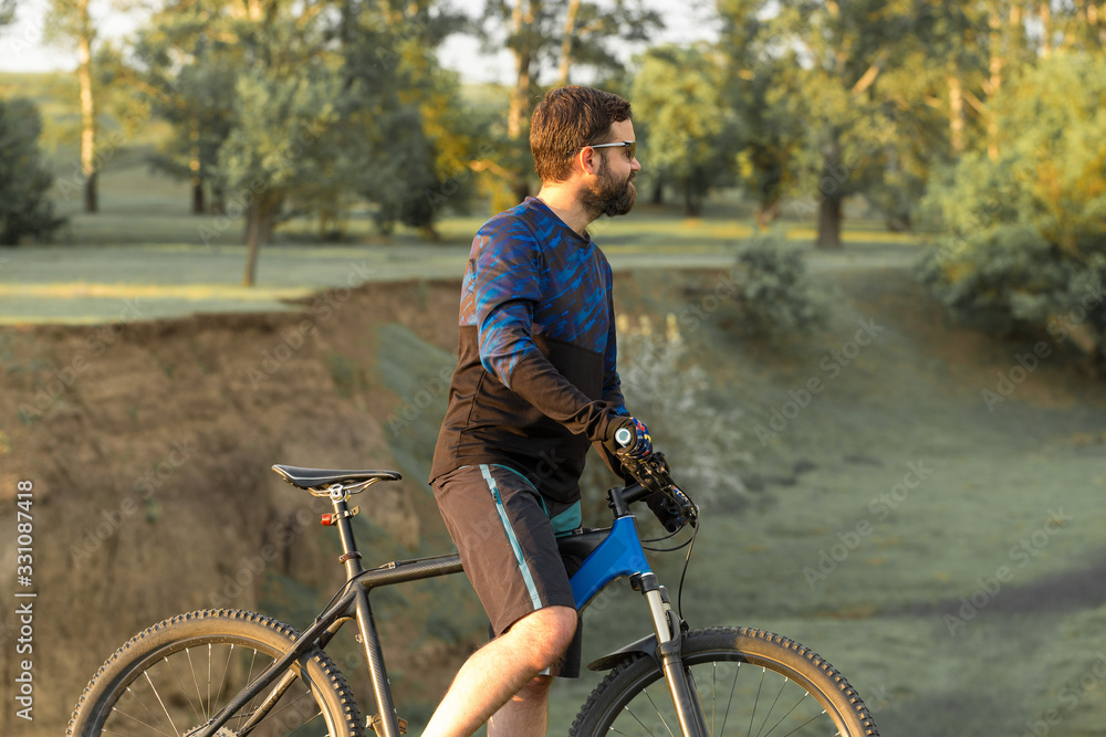 Cyclist in shorts and jersey on a modern carbon hardtail bike with an air suspension fork standing on a cliff against the background of fresh green summer forest Cyclist in shorts and jersey on a modern carbon hardtail bike with an air suspension fork standing on a cliff against the background of fresh green summer forest