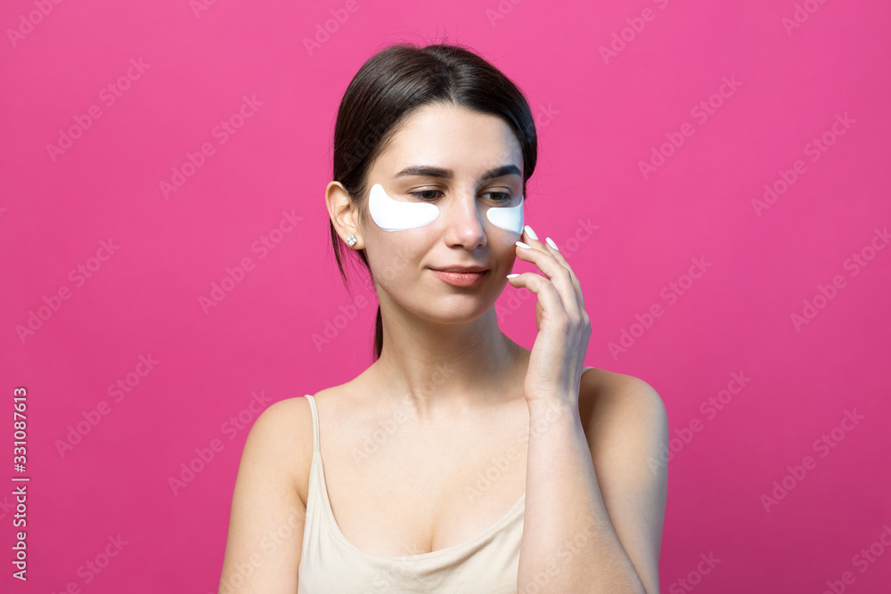 Close up portrait of pretty attractive girl with naked shoulders using patches under eyes. Standing over pink background. Close up portrait of pretty attractive girl with naked shoulders using patches under eyes. Standing over pink background.