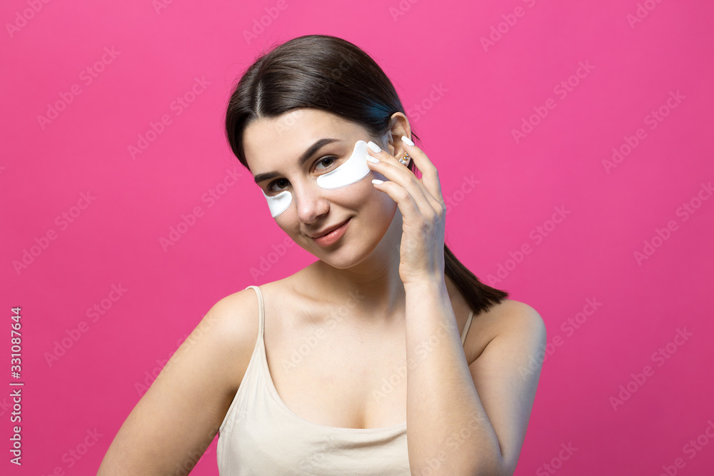 Close up portrait of pretty attractive girl with naked shoulders using patches under eyes. Standing over pink background. Close up portrait of pretty attractive girl with naked shoulders using patches under eyes. Standing over pink background.