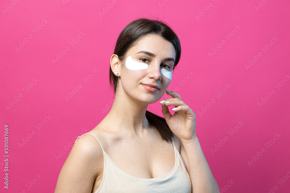 Close up portrait of pretty attractive girl with naked shoulders using patches under eyes. Standing over pink background. Close up portrait of pretty attractive girl with naked shoulders using patches under eyes. Standing over pink background.