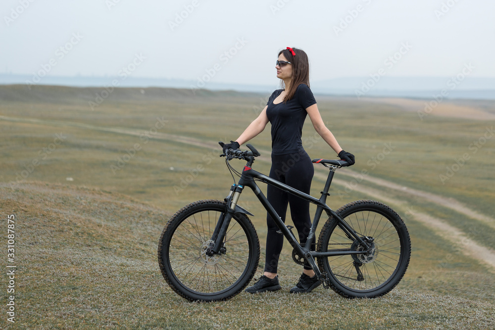 Fototapeta premium Girl on a mountain bike on offroad, beautiful portrait of a cyclist in rainy weather, Fitness girl rides a modern carbon fiber mountain bike in sportswear. Close-up portrait of a girl in red bandana.