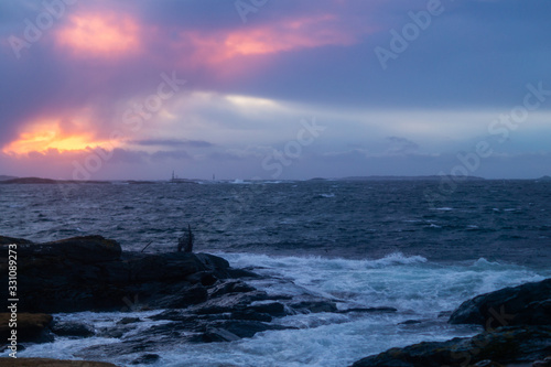 Sunset with waves hitting rocks on the shore line, clouds in the sky