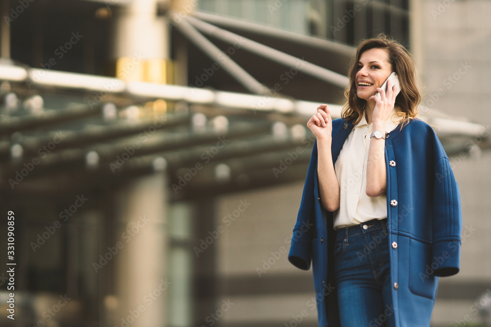 Fototapeta premium Caucasian business woman speaking by phone. Waist up portrait of a successful European business woman woman, talking on the phone, standing on glass background, modern office building. Sunny weather