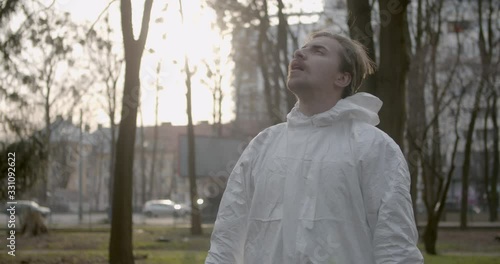 Portrait of male scientist in protective suit removing his mask and finnaly smelling fresh air in daytime park in pandemic coronavirus quarantine time as cars and pedestrians pass by 4K photo