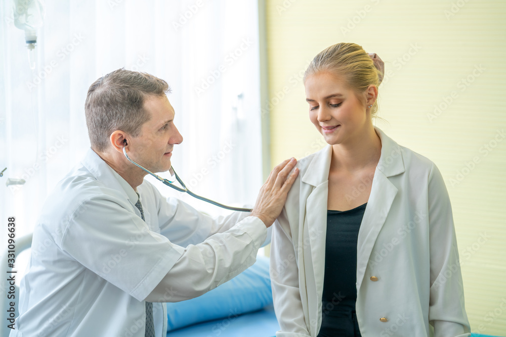 Obraz premium Mature Caucasian male doctor using stethoscope examining young beautiful woman patient in hospital clinic. Professional general practitioner consultant exam diagnosis medical healthcare concept