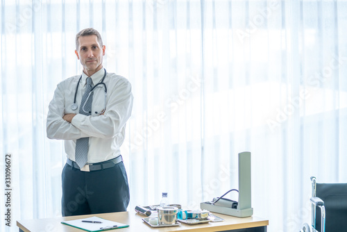 Foto Smiling gray hair mature male doctor standing in hospital examination room with looking at camera