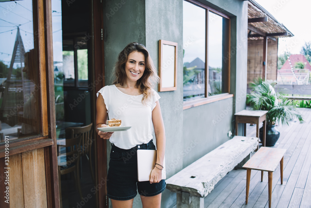 Smiling lady standing with piece of cake and laptop and looking at camera Smiling lady standing with piece of cake and laptop and looking at camera