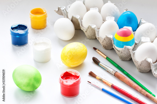 Painted Easter eggs and white eggs in a cardboard box, cans of paint, brushes on a white surface