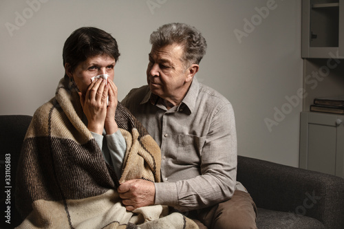 A man helps a sick woman on the sofa. A sick older woman sneezes into a napkin, and covered in a blanket. Family support for the illness. Coronavirus. COVID-19. A quarantine at home.