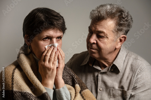 A man helps a sick woman on the sofa. A sick older woman sneezes into a napkin, and covered in a blanket. Family support for the illness. Coronavirus. COVID-19. A quarantine at home.