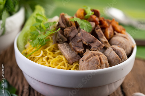 Yellow noodles in a cup with crispy pork, slices of pork, and meatballs together with Thai food-style noodles