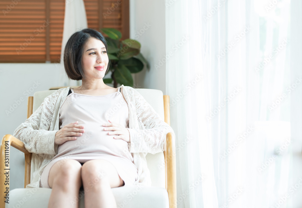 Happy Asian pregnant woman touching belly tummy with care on coach in bedroom look out window smiling, laughing. Enjoying pregnancy concept Happy Asian pregnant woman touching belly tummy with care on coach in bedroom look out window smiling, laughing. Enjoying pregnancy concept