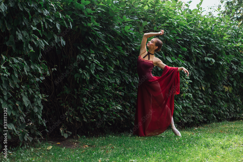 Woman ballerina in red ballet dress dancing in pointe shoes posing next to the wall of bushes