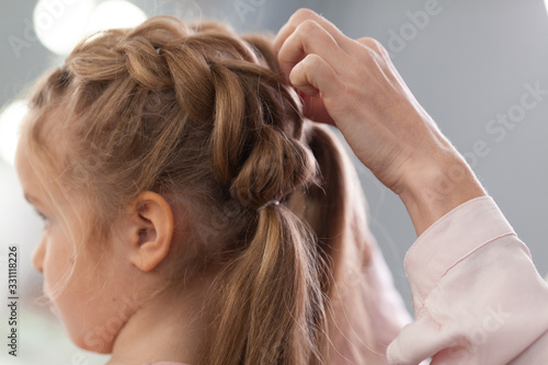 The work of the stylist on hairstyles in a bright salon, the girl makes curls and braids braids of a small beautiful model,