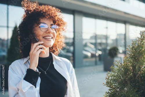 Lovely caucasian freelancer with curly hair is having a phone discussion while walking outside photo