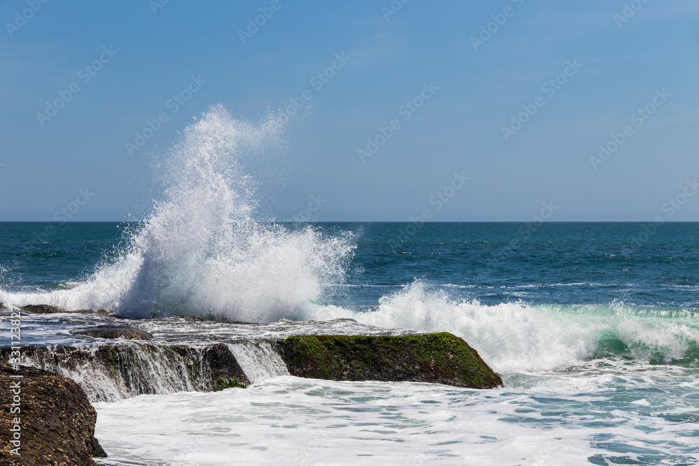 Wave breaking on rock during low tide near Tanah Lot Temple, Bali, Indonesia. Deep blue ocean and sky in the background. Wave breaking on rock during low tide near Tanah Lot Temple, Bali, Indonesia. Deep blue ocean and sky in the background.
