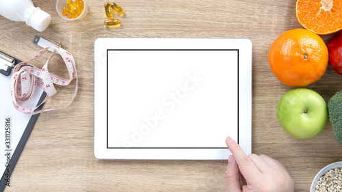 Nutritionist doctor hand pointing at digital tablet with blank screen for text, fresh fruits, vegetables, measuring tape and clipboard on wooden table. Healthy diet, nutrition and weight loss concept.