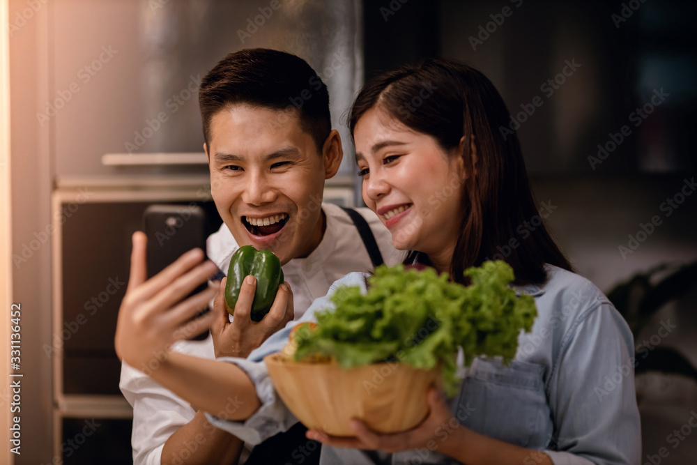 a Smiling Couple of Male and Female Using Phone in the Kitchen. Taking Selfie or Live Streaming via Smartphone while  Preparing a Vegan Food. Modern Healthy Lifestyle Concept. Cooking At Home