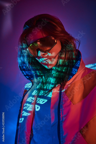 Attractive futuristic model wearing holographic glasses and racing blazer, posing in vivid neon studio lights. Cyberpunk concept