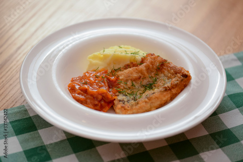 Fried fish filet with mashed potatoes and tomato sauce served on white plate on wooden table.