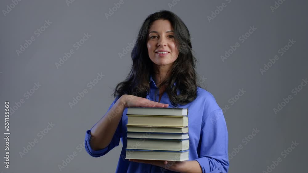 European smiling mature woman as student on grey background in studio holds stack of university books from library. Adult lady smiles, she is happy to graduate. European smiling mature woman as student on grey background in studio holds stack of university books from library. Adult lady smiles, she is happy to graduate.