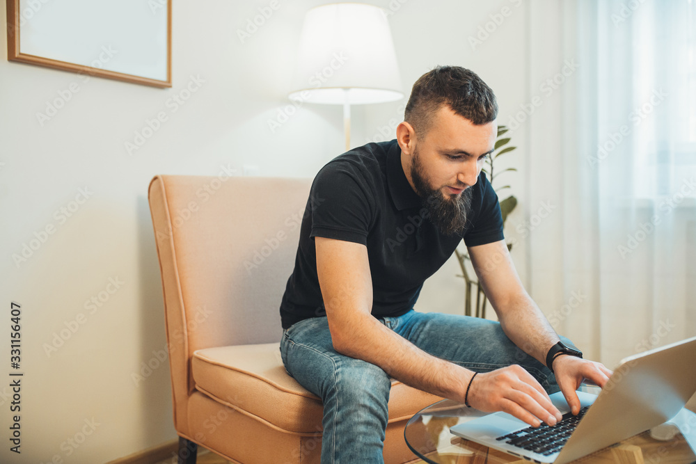 Bearded caucasian man with black hair working at home with computer while sitting on the armchair