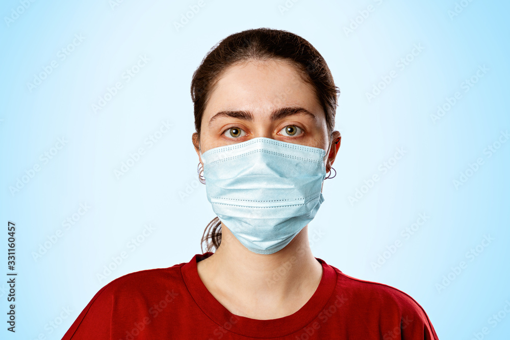 Portrait of a brunette woman in a medical mask and a red t-shirt, on a light blue background. Copy space. Concept of pandemic and protection against viral infection Portrait of a brunette woman in a medical mask and a red t-shirt, on a light blue background. Copy space. Concept of pandemic and protection against viral infection